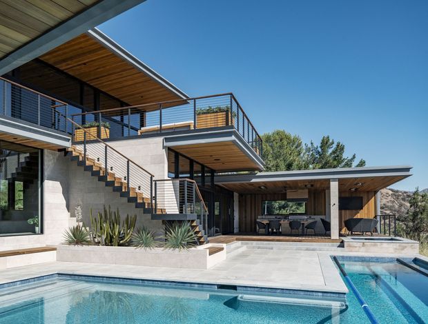 A contemporary multi-level residential exterior centered on an outdoor entertaining area and swimming pool. The house combines pale masonry walls, dark metal-framed glazing and railings, warm timber soffits, projecting balconies, and broad flat roof planes. An open-sided covered patio contains an outdoor dining and lounge area, while stairs connect the landscaped upper and lower terraces. The image is brightly lit with a clear blue sky and a restrained modern material palette.