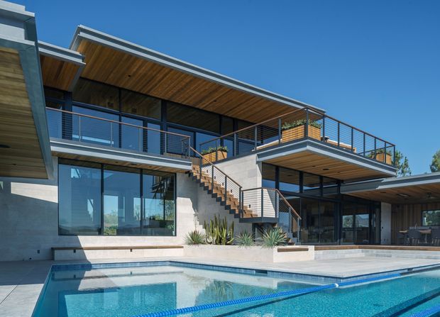 Contemporary multi-level residential home viewed from the pool, featuring broad floor-to-ceiling glazing, exposed concrete walls, timber-lined soffits, dark metal-framed windows, open-riser external stairs, and expansive upper and lower terraces overlooking a swimming pool.