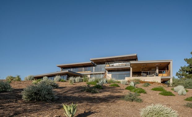 A wide exterior view of a contemporary multi-level residence set on a gently sloping, sparsely landscaped site. The home features layered low-pitched roof planes, extensive glazing, timber-toned soffits and cladding, and broad projecting terraces and balconies. Drought-tolerant planting and exposed earth surround the house beneath a clear blue sky.
