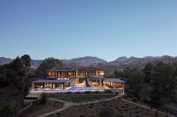 A large multi-level contemporary residence set into a landscaped, mountainous site. The building has extensive floor-to-ceiling glazing, horizontal flat roof forms, warm timber-toned illuminated interiors, broad terraces, and a long linear swimming pool. Curving paths and planted garden areas surround the house, with rugged mountains visible behind it.