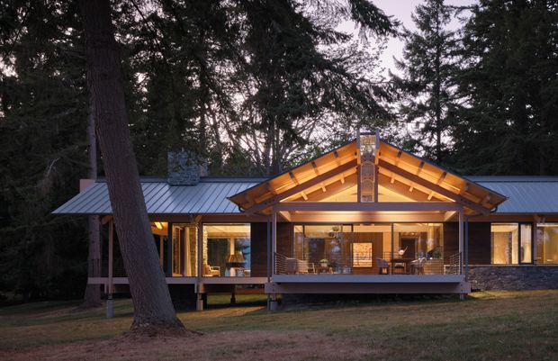 A single-storey forest residence viewed from the front at dusk. The home has a broad low-pitched metal roof, a prominent timber-framed gabled porch with exposed rafters, extensive warm-lit glazing, dark stone cladding, and a raised deck or entry platform. Tall trees surround the building, creating a secluded wooded setting.