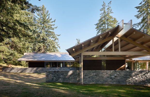 Exterior view of a low-rise, timber-framed building set within a wooded landscape. The composition features a broad asymmetric pitched roof with exposed rafters and structural beams, dark horizontal wall cladding, extensive glazing, and a substantial rough-stacked natural stone boundary wall. A secondary wing with a simpler metal roof extends to the left, while trees and lawn surround the structure.