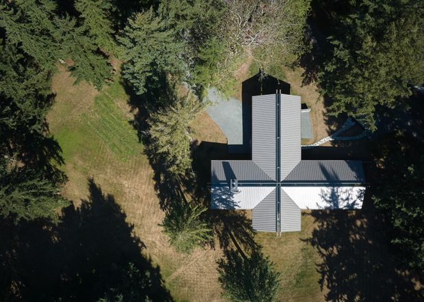 Aerial top-down view of a cross-shaped building surrounded by dense mature trees and lawn. The building has multiple intersecting pitched roof sections with dark grey corrugated metal roofing, creating a symmetrical radial plan. A pale gravel or concrete path is visible near the upper side of the building, while deep tree shadows cover parts of the site.