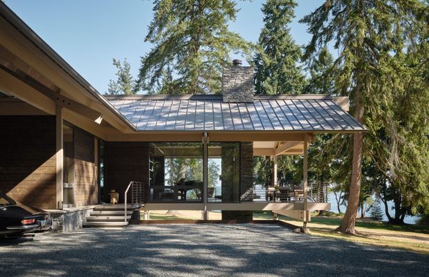 Exterior view of a low, contemporary timber-clad residence set among mature trees. The house has a broad metal roof with a central stone chimney, extensive floor-to-ceiling glazing, covered outdoor living areas, and a raised timber deck. A gravel driveway and carport-like covered parking occupy the foreground, with a glimpse of water or bright landscape beyond the trees.