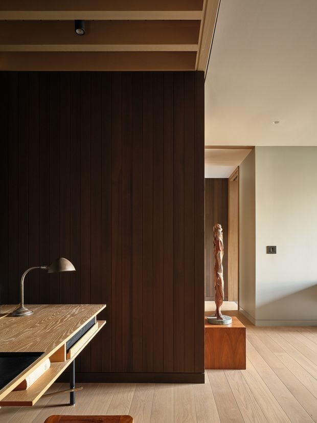 A refined contemporary interior corner featuring a dark vertical timber-panelled wall, integrated open shelving and a built-in timber desk or media console in the foreground. Warm light timber flooring, a sculptural artwork on a timber pedestal, recessed ceiling lighting and minimal cream-coloured walls create a calm, carefully detailed space.