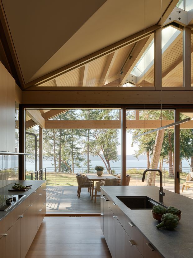A warm, light-filled contemporary kitchen opening onto a timber deck and landscaped waterfront setting. The room features a long island with an integrated stainless-steel sink and dark tapware, pale timber cabinetry, wood flooring, extensive dark-framed glazing, and a dramatic glazed view into the exposed timber roof structure above. A dining table sits beyond the kitchen, with water and trees visible through the windows.
