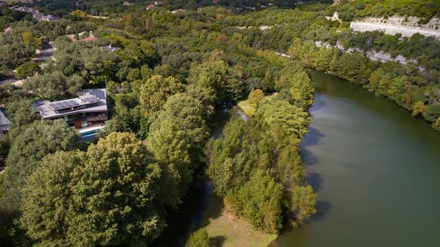 Aerial view of a modern, low-profile building nestled among dense mature vegetation beside a broad, calm body of water. The property includes dark roof surfaces and extensive glazing, with surrounding landscaped woodland and a nearby residential landscape visible in the distance.
