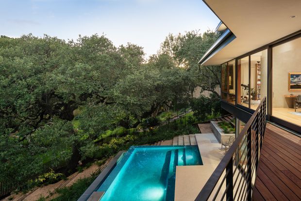 Exterior view of a contemporary multi-level home set among dense mature trees, showing a turquoise swimming pool, timber deck, concrete steps and terrace, dark metal balustrades, and extensive floor-to-ceiling glazing opening into a warmly lit interior. The building has a sharply angled roof overhang and a strong indoor-outdoor connection.