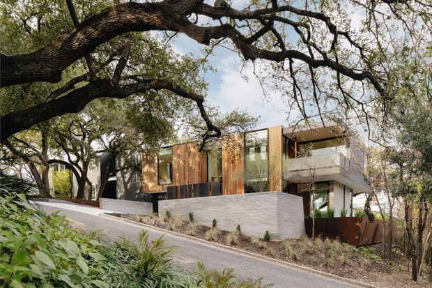 Exterior view of a contemporary multi-level residence set among mature trees on a sloping suburban site. The house features strong horizontal and rectilinear forms, extensive glazing, warm vertical timber cladding, pale masonry walls, projecting upper-floor volumes, and a dark metal entry or boundary element. A paved driveway or access road rises along the front, with landscaped planting around the building.