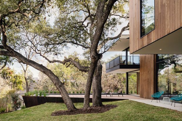 Exterior view of a contemporary multi-level residence integrated into a mature wooded landscape. The building features warm vertical timber cladding, extensive floor-to-ceiling glazing, slim dark metal framing, projecting balconies with glass balustrades, and a paved terrace with outdoor seating. Large established trees dominate the landscaped foreground and partially screen the architecture.