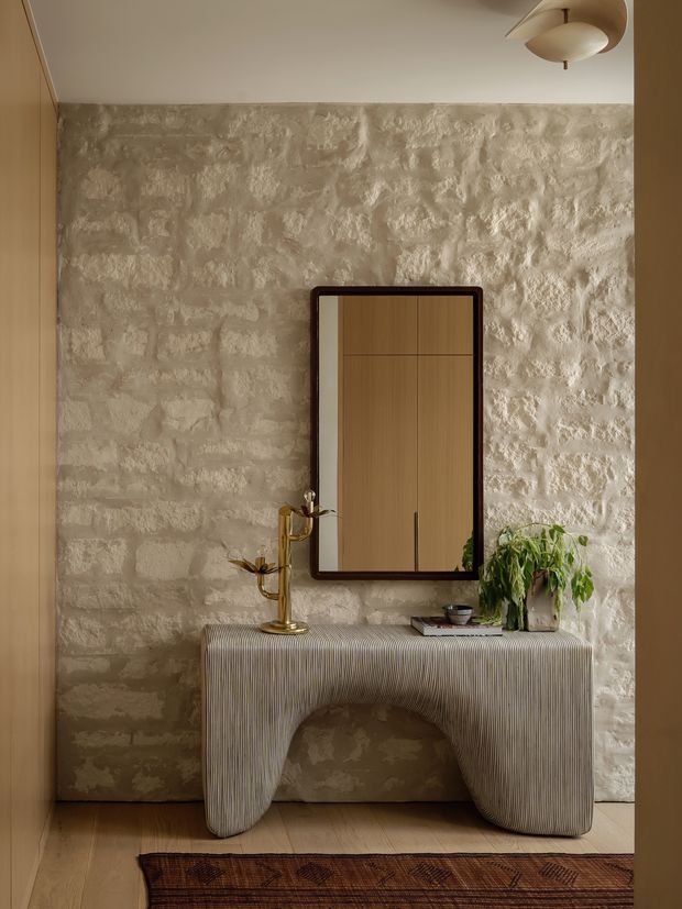 Warmly lit interior vignette featuring a sculptural ribbed console table against a rough textured stone or plaster feature wall. A dark-framed rectangular mirror, brass decorative object, books, and a potted plant sit on the console, with timber wall panelling at the sides, pale timber flooring, and a contemporary ceiling light. The styling is refined, earthy, and textural.