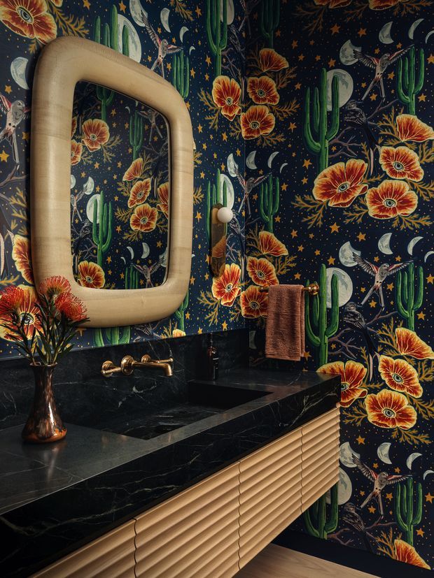 A richly styled bathroom vanity with a dramatic dark floral-and-cactus patterned wall covering, black marble benchtop and integrated basin, brushed brass tapware, a sculptural rounded rectangular mirror, and a fluted light-wood vanity front. The space has a compact, luxurious, highly decorative character.