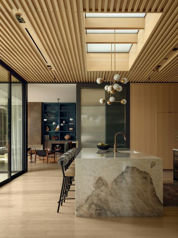 A refined contemporary kitchen opening into a living area, featuring a large veined natural-stone island with waterfall sides, warm timber cabinetry, dark-framed floor-to-ceiling glazing, and a dramatic timber slatted ceiling with a central skylight. A sculptural globe pendant hangs above the island, while built-in dark shelving and seating are visible beyond.