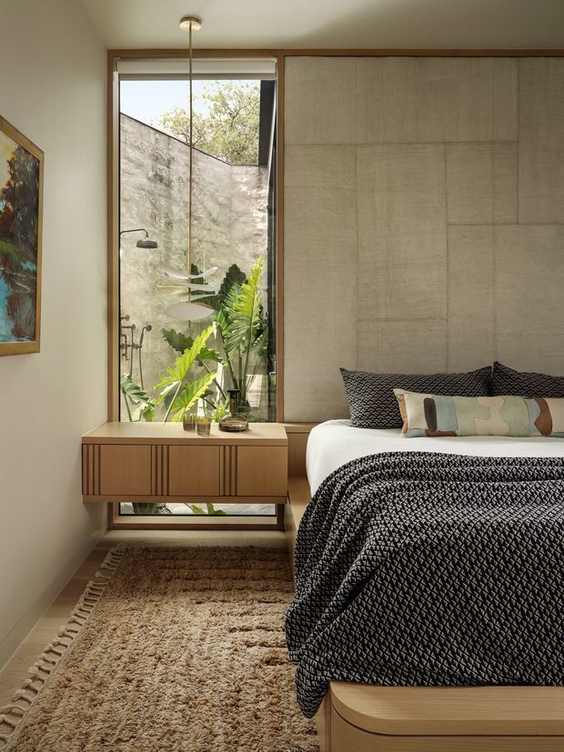 A warm, compact bedroom with a low platform bed dressed in a textured black-and-white quilt and patterned cushions. A timber wall-mounted console with vertical detailing sits beneath a tall window, which overlooks a private planted courtyard with a concrete wall. Natural timber trim, a beige textured rug, neutral walls, and a slim pendant light create a calm, refined interior.