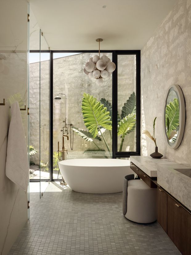 A refined contemporary bathroom with a freestanding white oval bathtub positioned beside a fully glazed shower enclosure. Large black-framed windows and doors open toward a planted courtyard, bringing in abundant natural light. The room features small-format pale grey mosaic floor tiles, textured light stone or plaster walls, a dark timber vanity with a pale stone benchtop, brushed metal plumbing fixtures, an oval wall mirror, and a sculptural clustered pendant light. The overall atmosphere is warm, serene, natural, and luxurious.