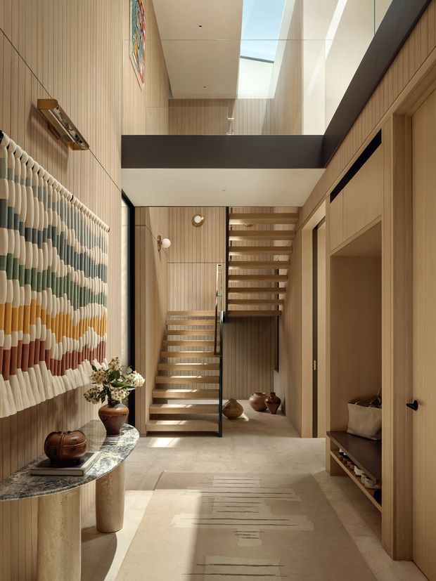 A refined double-height residential entry hall with an open staircase, warm vertical timber wall panelling, concealed built-in joinery, and a large linear skylight. A sculptural stone console with decorative pottery sits beneath a colourful textured wall hanging, while sunlight creates strong geometric patterns across the neutral floor and runner. The space has a calm, warm, contemporary character with a mezzanine or upper-level void visible above.