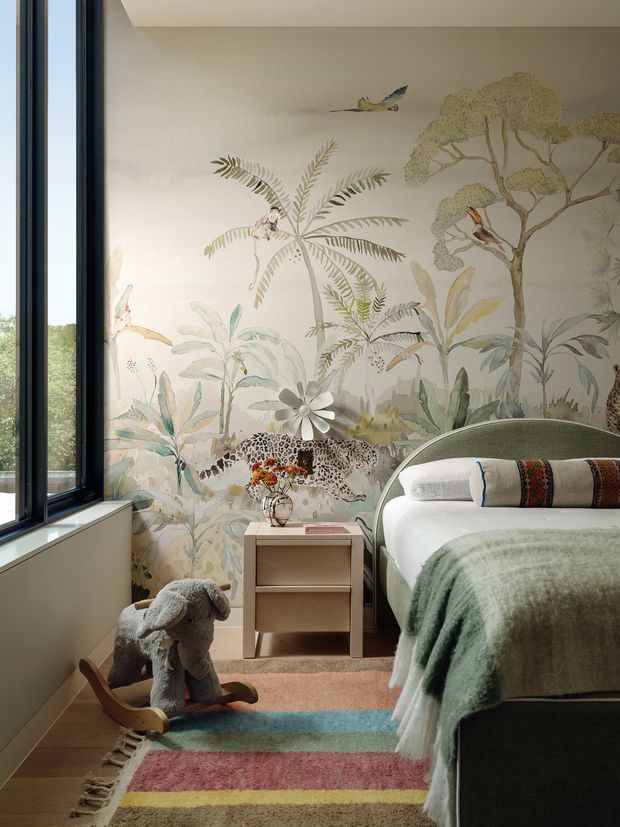 A playful, softly lit children's bedroom with a large illustrated jungle mural wallpaper, a curved upholstered bed, layered blankets and pillows, a compact bedside table, and a colourful striped rug. A rocking sheep toy adds a whimsical accent. Tall black-framed glazing along the left side brings in natural light and views of greenery.
