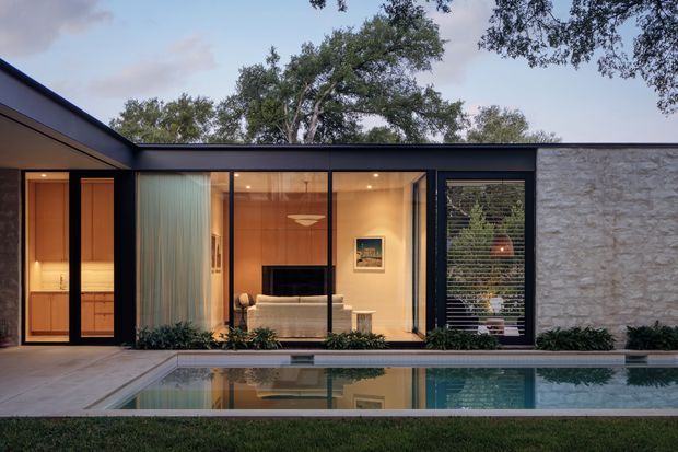Single-storey modern residence viewed from the pool courtyard at dusk, with a dark low-pitched roofline, extensive floor-to-ceiling glazing, black-framed doors and windows, and a light textured masonry wall. The glazing reveals a warm-toned living room with timber wall panelling, a sofa, television, and sculptural pendant light. A long reflecting swimming pool, pale paving, and low planting occupy the foreground.