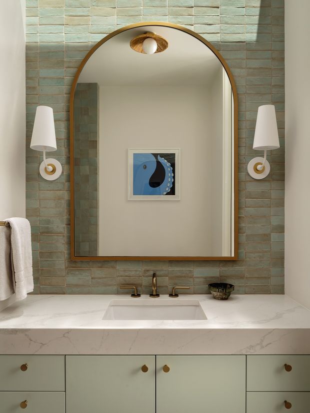 A refined bathroom vanity viewed straight on, featuring a large arched mirror with a warm bronze-toned frame, pale green textured rectangular wall tiles, sculptural wall sconces, a white integrated basin set into a pale stone benchtop, and sage-green cabinetry with small brass-toned pulls. A framed blue artwork is reflected in the mirror, adding a subtle accent.