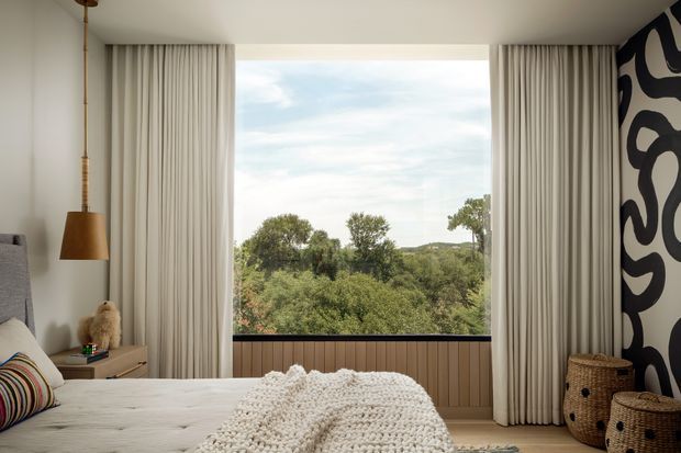 A contemporary bedroom with a large picture window overlooking leafy greenery. The room features floor-to-ceiling cream curtains, a low timber-panelled window seat or wall base, a white bed with a textured knitted throw, a striped accent cushion, a warm pendant light, woven baskets, and a bold black-and-white abstract feature wall.