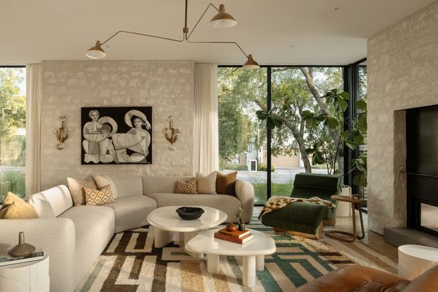 A warm, refined living room with a large cream sectional sofa, sculptural white coffee tables, patterned area rug, and a green upholstered lounge chair. Textured light plaster walls frame a black fireplace and a monochrome artwork, while oversized black-framed glazing opens the room to leafy outdoor views. Brass-toned sculptural wall lights and articulated ceiling pendants add character.