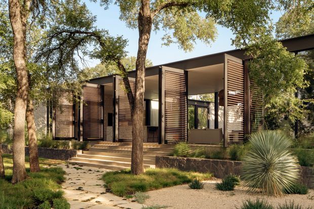 A low, modern pavilion-style house integrated into a wooded landscape. The facade uses extensive dark timber or metal horizontal louvres and screened openings beneath a broad flat roof, with large areas of glazing and sheltered outdoor living space. Stone retaining walls, broad steps, a pale irregular-stone path, and drought-tolerant planting connect the building to the site. Mature trees partially screen the structure.