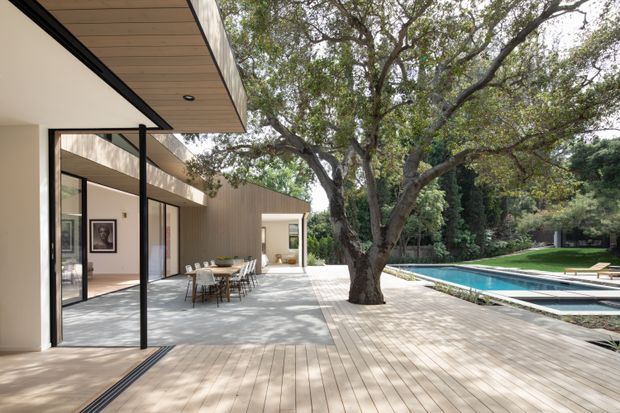 A contemporary single-storey residential outdoor entertaining area beside a long rectangular swimming pool. The covered patio has large sliding glass doors, slim dark frames, pale walls, a timber-lined soffit, and a dining setting. A broad timber deck surrounds a mature tree and extends alongside the pool, with landscaped garden beyond.