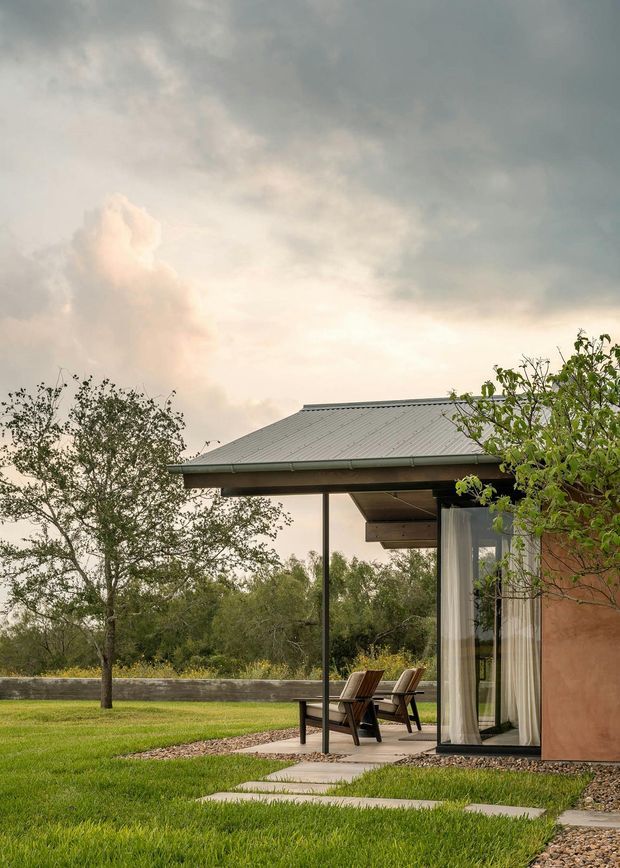 A contemporary single-storey pavilion-like residence set within a landscaped garden. The image focuses on a low-pitched grey metal roof with broad eaves, slender dark supports, large glazed sliding doors with sheer curtains, and a warm earth-toned exterior wall. Two outdoor lounge chairs sit on a paved terrace beside a manicured lawn, gravel beds, and stepping-stone paths. The scene has soft overcast evening light and a calm, natural atmosphere.