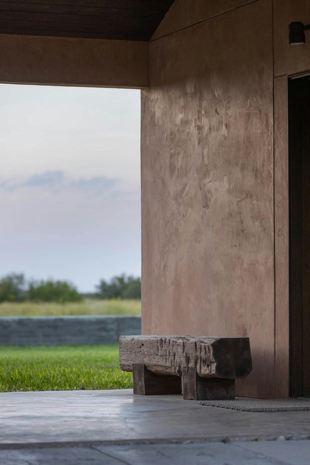 A moody architectural threshold or covered outdoor entry with a rough brown plaster wall, dark timber-lined ceiling, and a heavy rustic timber bench. The open side frames a grassy landscape and low dark boundary wall. The floor appears to be large smooth grey concrete or stone slabs, and a dark doorway is visible at right.