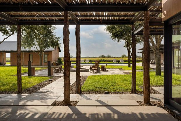 A covered outdoor terrace or walkway framed by rough timber posts and beams beneath a slatted timber roof. Concrete paving leads through landscaped lawns toward a seating area and low rectangular reflecting pool or water feature, with a simple low-rise building visible beyond. The scene has a warm, rustic-modern architectural character and strong indoor-outdoor connection.