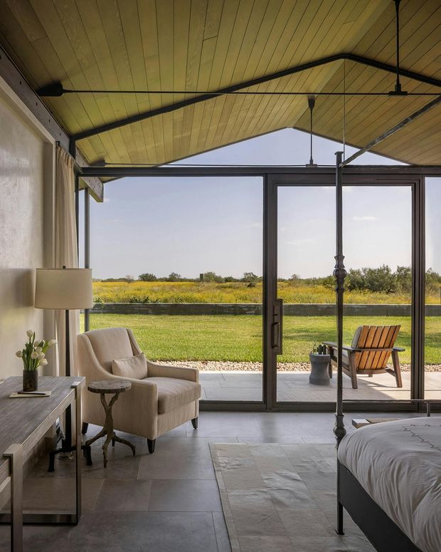 A calm, light-filled bedroom with expansive dark-framed sliding glass doors opening to a lawn and rural landscape. The room features a timber-lined angled ceiling with exposed dark metal structural bracing, pale tiled flooring, a neutral upholstered armchair, slim console table, bedside-style lamp, and a partially visible contemporary bed. An outdoor chair and patio are visible beyond the glazing.