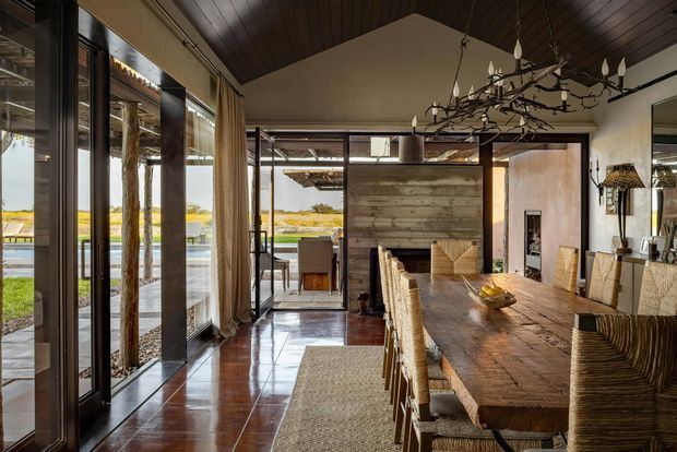 A warm, rustic dining area with a long solid timber dining table and woven dining chairs. The room features polished dark floor tiles, a dark timber-paneled pitched ceiling, full-height glazed doors with curtains, and a timber-and-glass opening toward an outdoor entertaining area. A sculptural branch-like chandelier hangs above the table, while a weathered timber feature wall and natural woven textures reinforce the earthy character. The landscape is visible through the glazing.