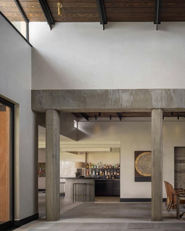 Interior view of a refined modern space with exposed concrete columns and beam, dark timber ceiling panels with black structural beams, pale plaster walls, and large grey floor tiles. A compact dark home bar and kitchen joinery are visible beyond the opening, with a dining area and timber-framed glazing at the side. The composition highlights layered circulation, structural detailing, and a warm industrial material palette.