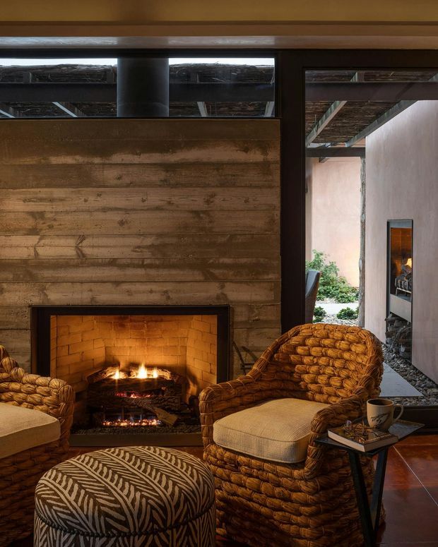 A cozy rustic living room centered on an open brick fireplace set into a horizontally timber-lined feature wall. Woven rattan lounge chairs and a matching patterned ottoman frame the fire, while a dark-framed glazed opening reveals an adjoining outdoor courtyard and covered timber structure. Warm low lighting, earthy brown tones, and natural textures create an intimate retreat-like atmosphere.