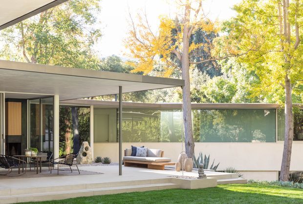 A modern single-storey home opening onto a landscaped courtyard or terrace, with broad flat roof planes, slender steel supports, extensive glazing, and pale rendered walls. Outdoor seating, a low built-in bench, sculptural planters and drought-tolerant plantings sit beneath mature trees. The scene has a bright, calm mid-century character with strong indoor-outdoor connection.