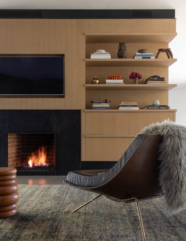 A refined living room centered on a built-in timber entertainment wall and open shelving. The composition includes a wall-mounted television, black stone or dark marble fireplace surround with an active fire, carefully styled shelving, and a sculptural dark leather lounge chair with a sheepskin throw. The room has a warm, moody, sophisticated character with layered natural textures.