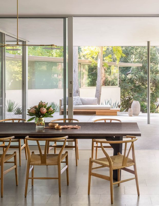 Bright, open-plan dining area with a long dark timber dining table and woven-seat timber chairs. Floor-to-ceiling glazed doors and windows open onto a covered outdoor lounge or terrace, with a low built-in bench, greenery, and sculptural planters visible beyond. The interior has pale tiled flooring, a light ceiling, slim neutral frames, and a linear brass-toned pendant light. The overall look is calm, refined, and nature-connected.