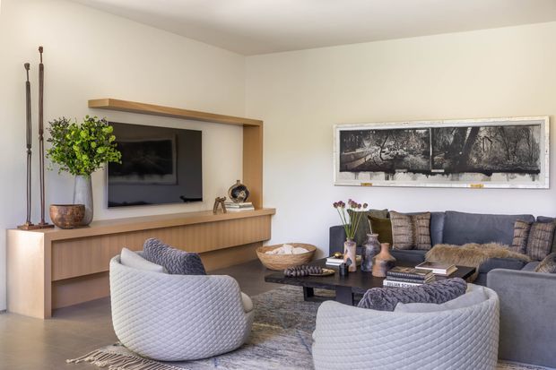 A warm contemporary living room with a built-in timber entertainment wall and television, two rounded upholstered lounge chairs, a large sectional sofa, a low dark coffee table, textured rug, and neutral decorative styling. A large framed monochrome artwork hangs above the seating area.