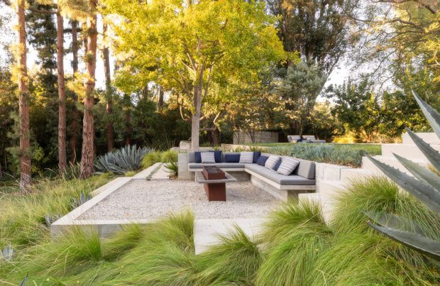 A landscaped outdoor lounge area set within a mature, wooded garden. A built-in L-shaped light-grey sectional surrounds a low dark coffee table on a pale gravel terrace, with raised concrete planters, ornamental grasses, agave-like plants, and stepped garden levels creating a natural yet refined setting.