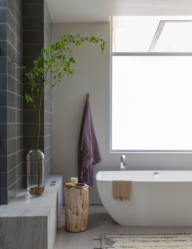 A bright, refined bathroom featuring a freestanding white bathtub beneath a large skylight and window. The space combines dark charcoal rectangular wall tiles with pale grey walls and marble-look stone surfaces. A simple chrome wall-mounted tap, timber stool, glass vase with greenery, towels, and a neutral textured rug add natural accents.