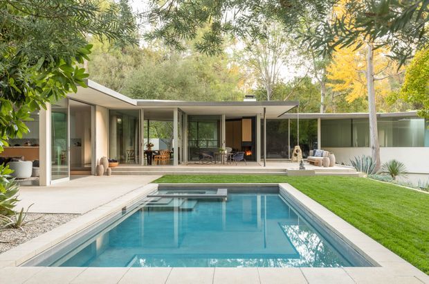 A low-profile modern home opens onto a landscaped backyard with a rectangular swimming pool in the foreground. Extensive floor-to-ceiling glazing, slender structural posts, flat roof planes, pale paving, and a covered outdoor living area create a seamless indoor-outdoor composition. Mature trees, lawn, and drought-tolerant planting surround the house.