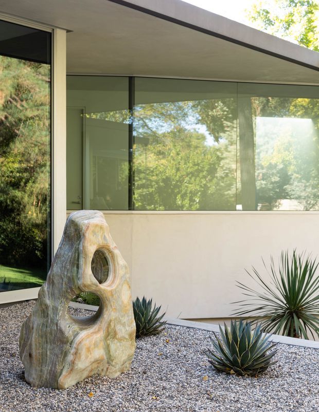 A minimalist modern exterior courtyard or landscaped entry area beside a low, flat-roofed building. Large dark-framed glazed openings reflect surrounding trees, while a pale rendered wall and deep roof overhang create a clean horizontal composition. The foreground is covered in light gravel with sculptural stone and drought-tolerant agave/yucca planting.