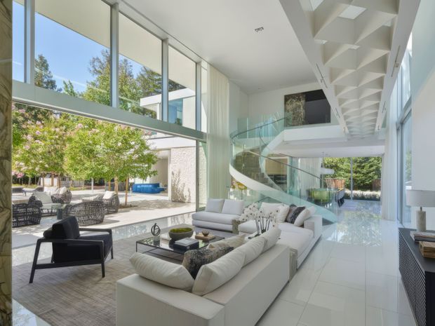A bright, expansive contemporary living room with floor-to-ceiling glazing opening onto a landscaped courtyard and pool area. White modular sofas and dark lounge chairs surround a low glass coffee table on pale flooring. A sculptural curved staircase with glass balustrades leads to a mezzanine, while a dramatic geometric coffered ceiling and double-height glazing emphasize the scale and openness of the space.