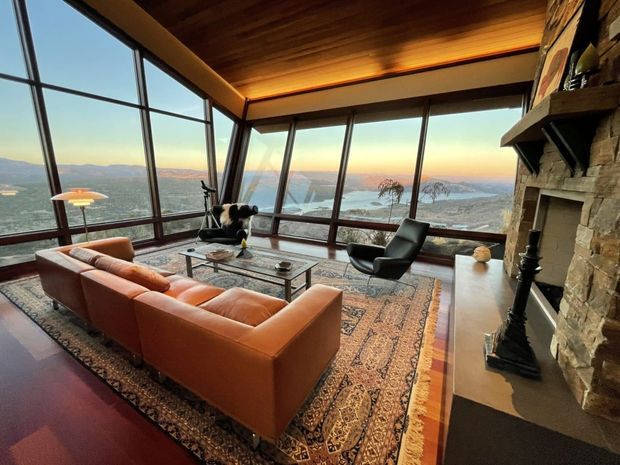 A warm, mid-century-inspired living room with a large rust-orange sectional sofa, black lounge chair, glass coffee table, patterned area rug, and stone fireplace. The room features extensive angled floor-to-ceiling glazing overlooking a mountainous or coastal landscape, with a rich timber ceiling and dark window frames.