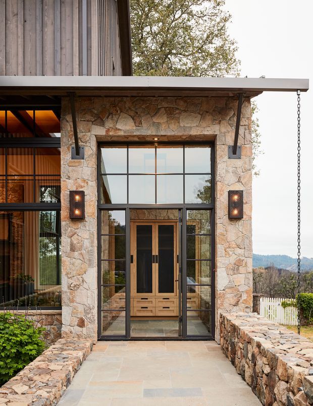 A close exterior view of a contemporary residential entryway beneath a broad flat canopy. The entrance is framed by irregular natural stone masonry, tall black-framed glazed doors and sidelights, and a timber double door with integrated drawers or joinery visible behind the glazing. A chain rainwater downspout hangs at the canopy edge, while stone boundary walls and a landscaped rural or wooded setting surround the approach.