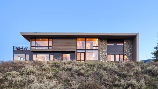 A contemporary two-storey residential house viewed from the front across native scrubland. The building has a strong rectilinear form with a broad flat roof, extensive dark-framed glazing, horizontal timber cladding, and a central section of irregular grey stone veneer. A rooftop or upper-level terrace with glass balustrading is visible on the left. The surrounding vegetation and open blue sky emphasize the building's connection to a natural, elevated landscape.