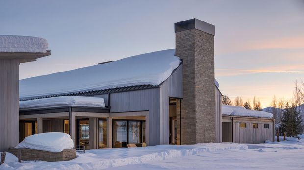Exterior view of a contemporary single-storey building in a snowy landscape, featuring a broad low-pitched roof heavily covered in snow, vertical grey cladding, extensive timber-framed glazing, and a tall rectangular stone-clad chimney or fireplace volume. The scene is photographed at dusk with snow-covered outdoor areas and trees in the background.