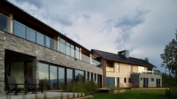 Exterior view of a contemporary multi-level house on a landscaped site. The facade combines extensive stacked natural stone masonry with warm vertical timber cladding, dark pitched roof forms, large glazed openings, and a glass-balustraded upper terrace. A recessed outdoor seating area and lawn occupy the foreground, with distant mountainous terrain visible through the glazing and beyond the property.