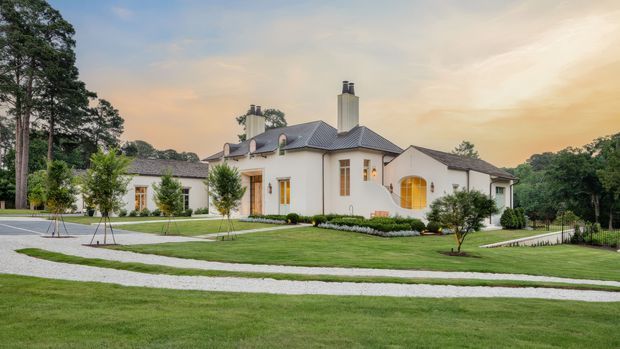 A large single-storey residential estate set within expansive landscaped grounds. The pale rendered exterior has a symmetrical, traditional composition with multiple wings, dark hipped tiled roofs, tall chimneys, timber-framed windows, and a prominent arched glazed entrance. Curving pale gravel or paved walkways cross the manicured lawn, with mature trees and newly planted landscaping surrounding the house.