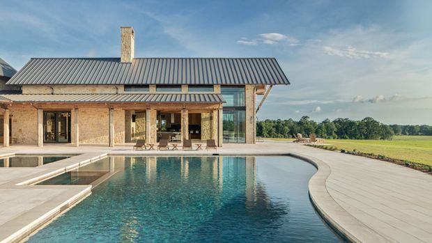 Exterior view of a substantial brick-clad house with a long corrugated metal roof, covered outdoor terrace, extensive glazed corner, and a large curved swimming pool in the foreground. The property opens onto a broad rural landscape and appears to be a single-storey, pavilion-like residence with strong indoor-outdoor connection.